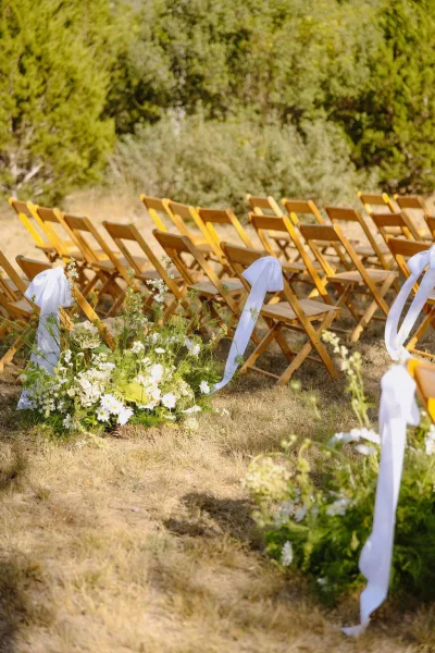 Ceremony aisle decor for outdoor ceremony seating features white and green florals edging a grass lawn amid garden shrubs and trees.