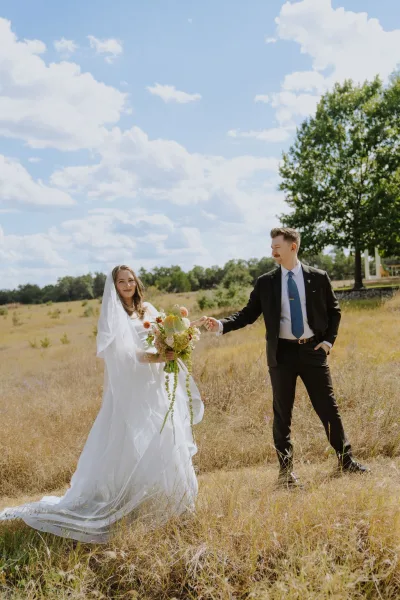 Couple portrait of a bride and groom holding hands in a sunlit grass field, her bridal bouquet bright against trees and a blue sky.