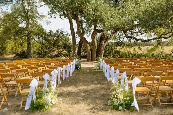 Outdoor ceremony setup features wooden ceremony chairs tied with white ribbons lining a natural aisle beneath oak trees in a dry, open field.