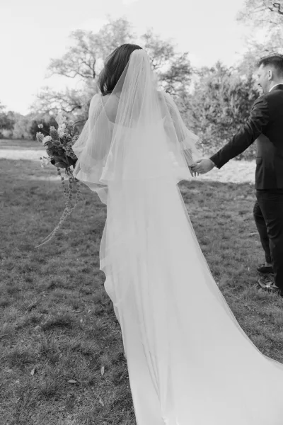 Couple portrait of bride and groom holding hands in an open field, her veil trailing as they walk across the grass with trees under a wide sky.