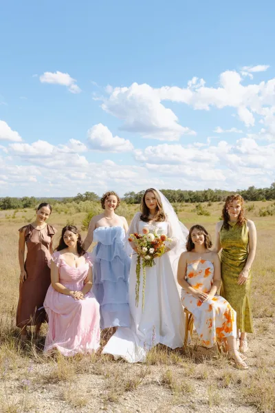 Bridal party portrait of a bride with bridesmaids in an open field under a blue sky, the bride holding a lush bouquet as clouds drift behind.