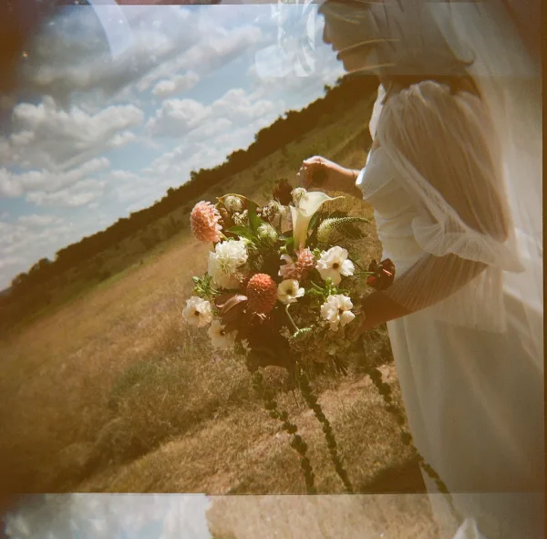 Bridal bouquet, a peach and ivory bouquet with trailing greenery, shown in a grassy field under a cloudy sky with a soft tree line.