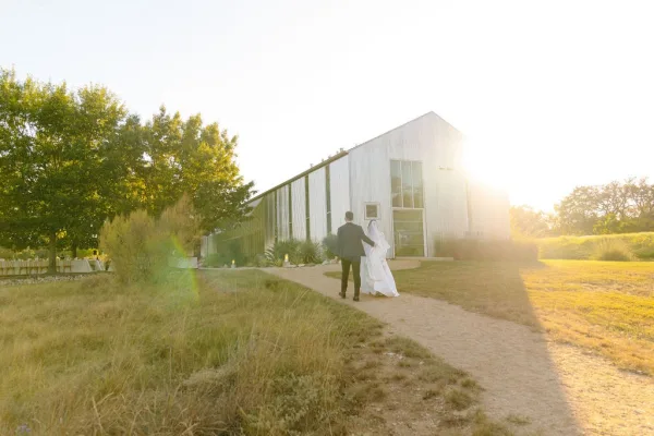 Couple portrait of bride and groom walking hand in hand beside a barn along a sunlit gravel path, her veil catching the light.