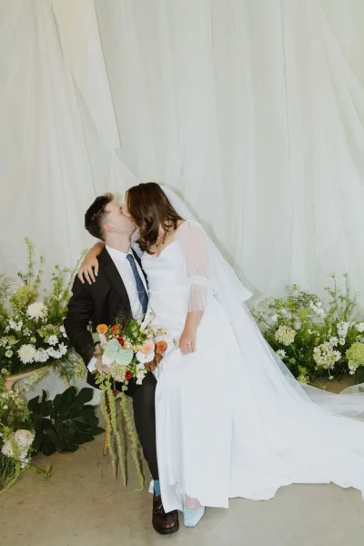 Wedding kiss portrait with bride on groom's lap, her cascading greenery bouquet against a draped fabric backdrop and surrounding greenery.