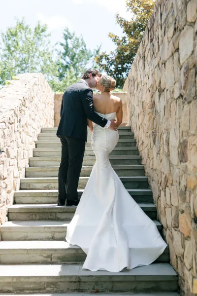 Wedding kiss portrait of a bride and groom on stairs, her gown train sweeping a sunlit stone staircase framed by greenery and trees.