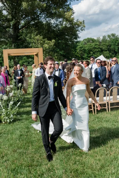 Wedding recessional with a just married exit as the couple walks hand in hand down the aisle, framed by a wooden ceremony arch on a grass lawn
