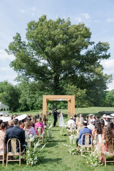 Ceremony moment at an outdoor wedding ceremony as the couple stands beneath a wooden arch under a large tree on a sunny lawn.