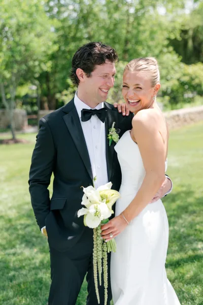 Couple portrait of bride and groom laughing as she holds white calla lilies on a garden lawn beside a stone wall and trees.