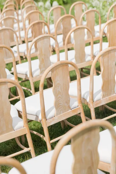 Ceremony seating features outdoor ceremony chairs with white cushions on a grass lawn bordered by greenery and white flowers.
