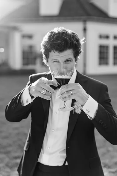 Groom portrait as he lifts a coupe glass, smiling on a grass lawn near a building exterior with windows.