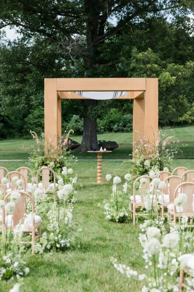 Ceremony setup for outdoor ceremony decor with a wood ceremony arch on a grass lawn beneath a large tree and surrounding garden greenery.
