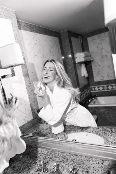 Bride getting ready with soft bridal makeup, touching up lipstick in a marble-tiled bathroom with granite countertop and warm sconce light.