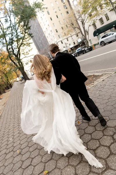 Couple portrait of the bride and groom walking hand in hand along a tree-lined city street, her flowing dress train trailing behind.
