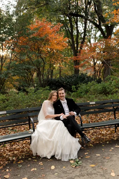 Couple portrait of bride and groom sitting on a park bench, a fall wedding portrait framed by golden autumn trees and crisp fallen leaves.