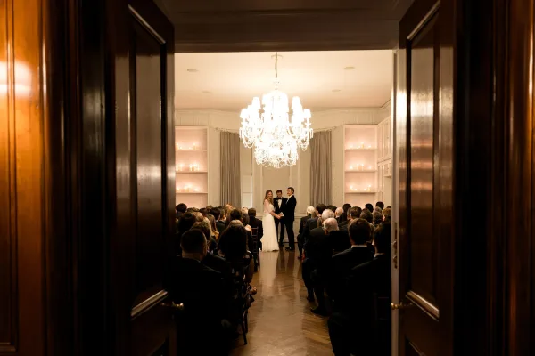 Ceremony moment during an indoor wedding ceremony as the bride and groom hold hands beneath a glowing chandelier.