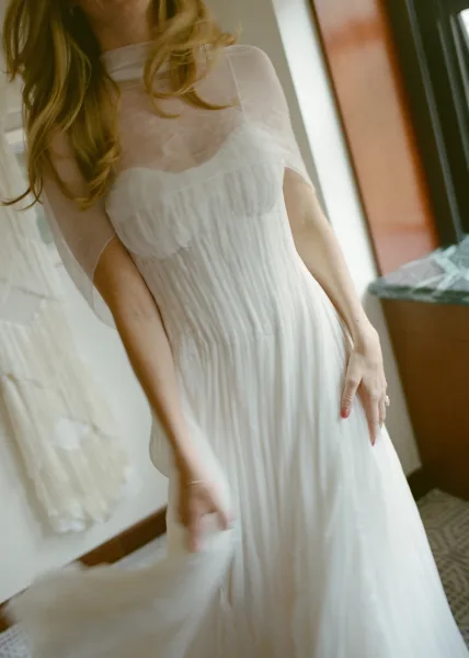 Wedding dress close-up featuring a sheer tulle cape drifting over an ivory gown, captured near a patterned floor tile and wood-trimmed window.