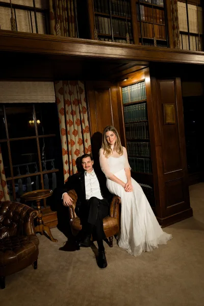 Couple portrait of a bride and groom in a library, groom seated in a leather armchair, bride standing by bookshelves and wood paneling.