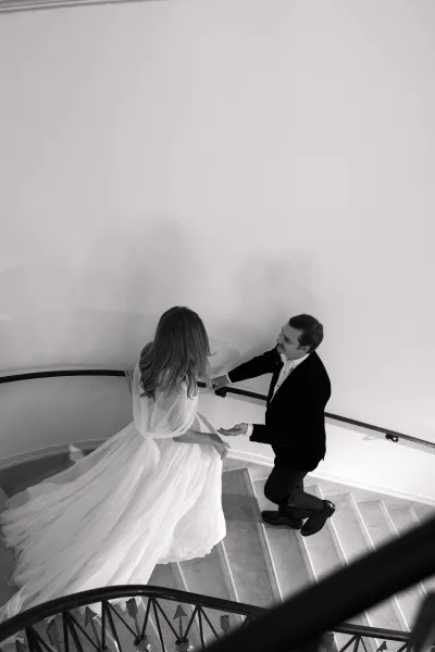 Couple portrait of a bride walking down stairs with a long train, groom by her side on a curved marble staircase against a bright white wall.