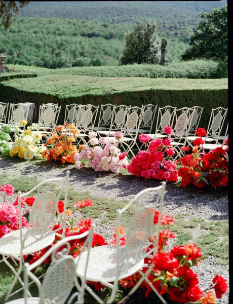 Ceremony aisle design with white metal chairs flanking a gravel path, as outdoor ceremony decor framed by garden hedges and distant hills.