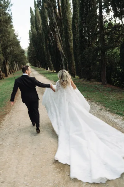 Bride and groom walking in a couple portrait along a tree-lined gravel path, her lace cape trailing as he matches pace beside her.