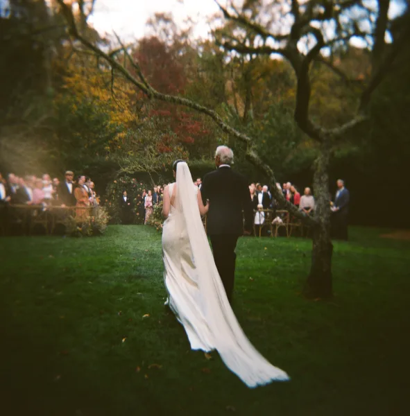 Wedding processional with bride walking down aisle in a long veil and train, escorted by her father on a garden lawn with guests standing