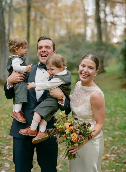 Wedding family portrait of bride and groom with kids, bride holding bouquet, standing in an autumn park with trees and fallen leaves