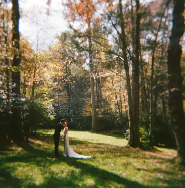 Wedding couple portrait of newlyweds kissing in sunlight among forest trees and autumn leaves, bride in long train dress with groom in tuxedo