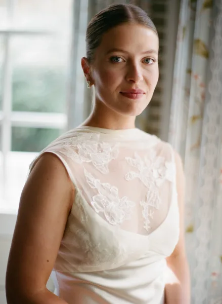 Bridal portrait of a bride by window in natural light, wearing a lace illusion neckline dress and pearl earrings with soft curtains behind