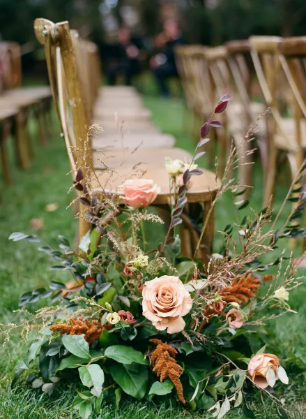 Ceremony aisle decor with a low peach rose and greenery floral cluster on grass beside wood folding chairs on a garden lawn