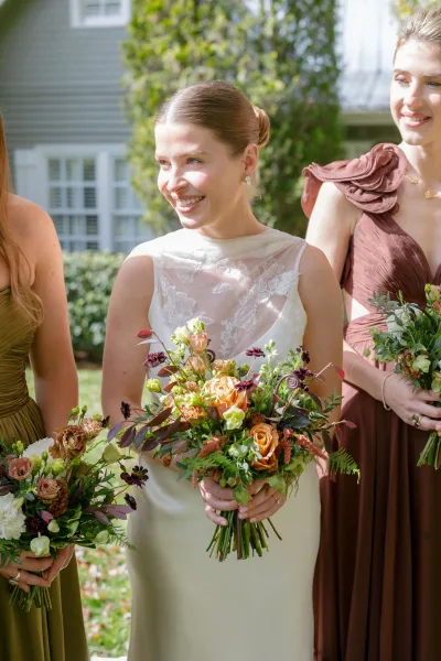 Bridesmaid portrait with bridesmaids holding bouquets in rust and olive dresses, orange rose and burgundy blooms, on a garden lawn near a house