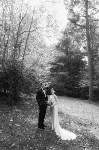 Couple portrait in a black and white wedding portrait style, bride in sleeveless gown holding bouquet beside groom in tuxedo on a woodland lawn