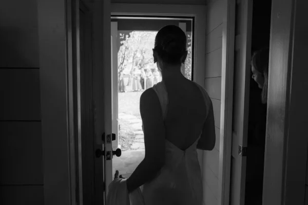 Bridal entrance as bride walks through doorway from behind in a backless wedding dress, hair bun and earrings, holding her train toward a tree-lined path