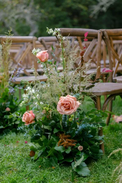 Ceremony aisle flowers with a low peach rose and greenery arrangement beside wooden chairs on a garden lawn under trees