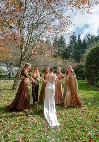 Bridesmaids with bride in a circle, holding bouquets with the bride in a white gown on a lawn with autumn trees and fallen leaves