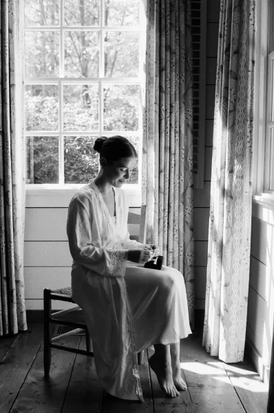 Bridal getting ready as the bride putting on jewelry in a lace-trim robe by a large window, ring box on chair, trees outside
