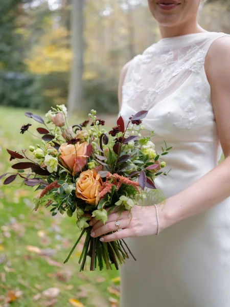 Bridal bouquet with fall wedding bouquet colors, rust orange roses and greenery held against a lace bodice with ring in a garden setting