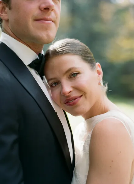 Couple portrait of bride leaning on groom, her head on his shoulder, in lace wedding dress beside tuxedoed groom in soft greenery