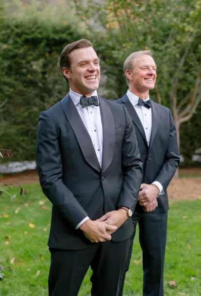 Groom portrait in a black tuxedo with bow tie and boutonniere, showing wristwatch and wedding band, standing on a garden lawn under trees