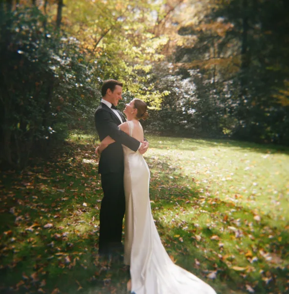 Couple portrait of bride and groom embrace, her satin gown train and his black bow tie, on a dappled garden lawn with fallen leaves