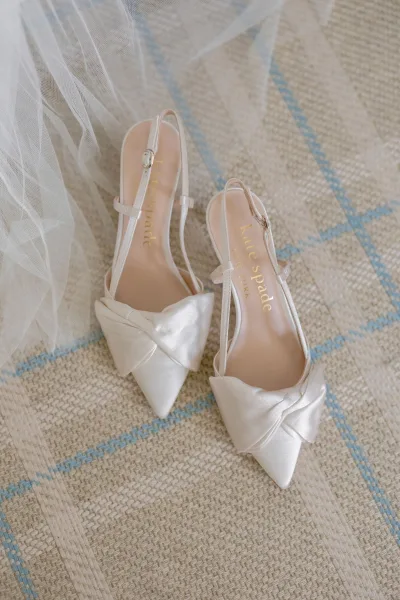 Bridal shoes in white satin with bow and ankle straps, styled with a tulle veil on a woven blue-striped rug flat lay