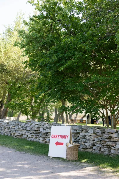 Wedding ceremony sign with ceremony direction sign arrow on an easel by a stone wall and gravel path, with a parasol basket nearby