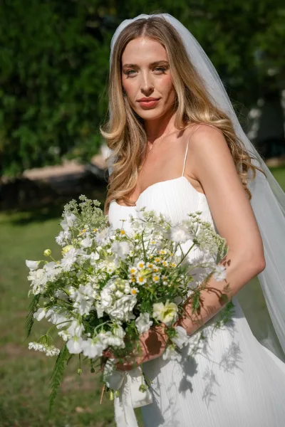 Bridal portrait of a bride holding bouquet in a white gown and long veil, sunlight filtering through green trees and lawn behind her