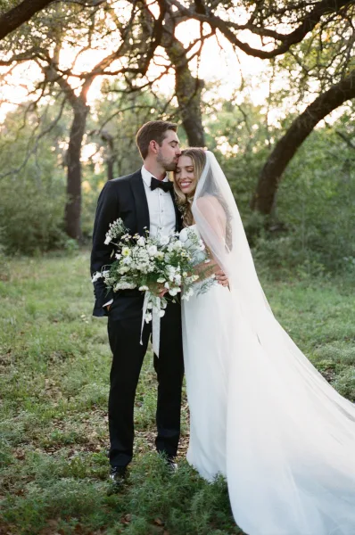 Couple portrait of groom kissing bride’s forehead as she holds a bouquet with veil flowing, framed by woodland trees in sunset light