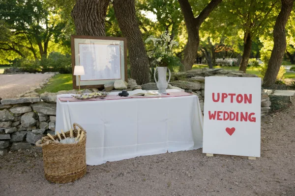 Wedding welcome table with a wedding welcome sign, framed seating chart, guest book, and white florals by a stone wall with string lights