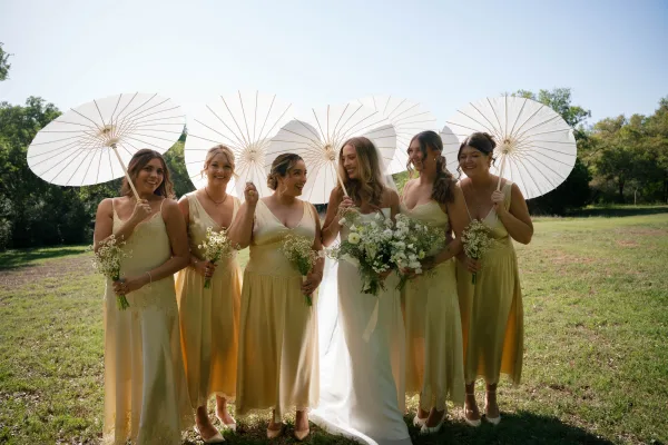 Bridesmaids portrait of the bride with bridesmaids holding white paper parasols and bouquets in a grassy field under blue sky