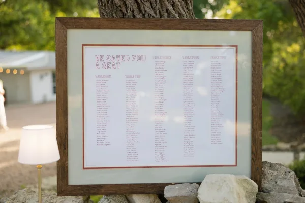 Wedding seating chart framed in a wooden frame with printed guest list and table assignments, displayed outdoors beside a tree trunk and lamp