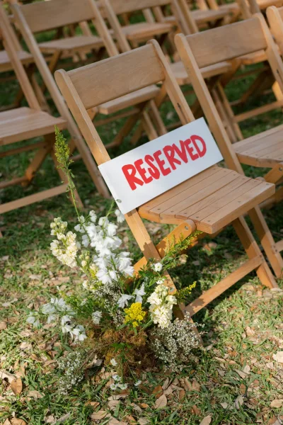 Reserved wedding seating with a reserved sign on wooden folding chairs, lined with aisle flowers and greenery on a leaf-strewn lawn