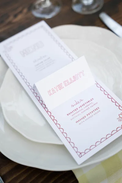Wedding menu card on a wood table reception place setting with white plates, silverware, glasses, and a printed place card accent