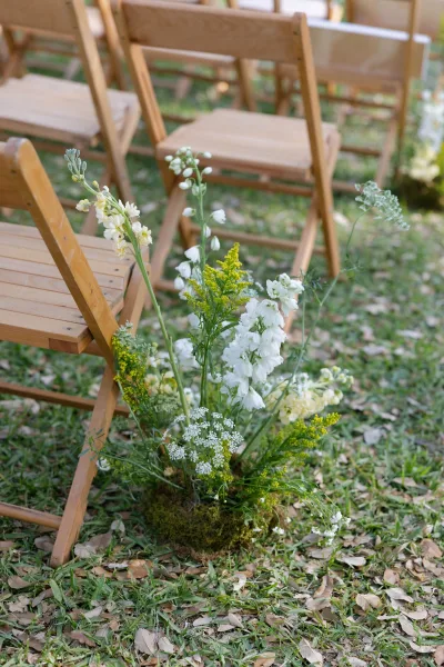 Ceremony aisle decor with an aisle flower arrangement of white and yellow blooms and greenery by wood folding chairs on a grassy lawn