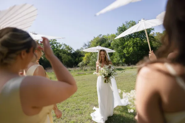 Bridal portrait of bride holding bouquet with veil, walking in a sunny meadow with bridesmaids in yellow dresses under white parasols